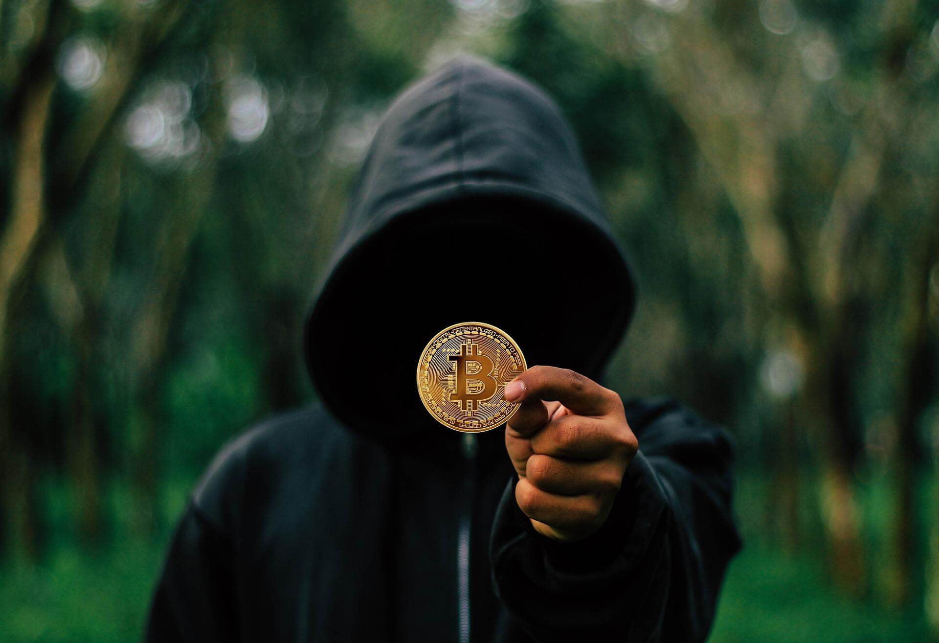 Persona con sudadera y capucha negra sosteniendo la representación visual de la criptomoneda bitcoin.