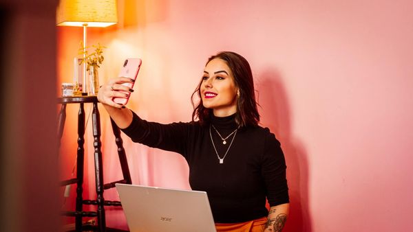 Mujer tomándose una selfie y trabajando en su computadora portátil en un escritorio con una pared rosa.